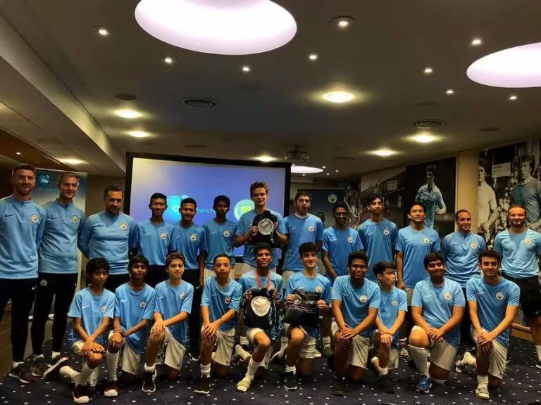 How The Qnet-Man City Football Partnership Empowers Young Talents From Around The World 3 Kids from Indonesia in Man City Etihad Campus locker room posing with coaches and players.
