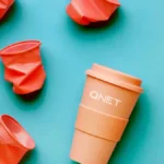 Orange reusable QNET tumbler surrounded by crumpled plastic cups